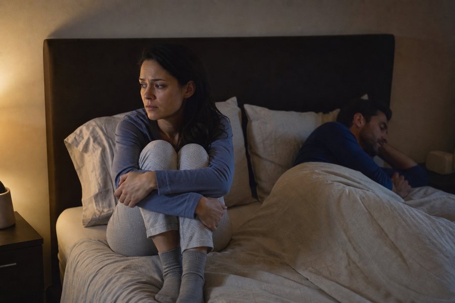 Woman sitting up awake and worried in bed while her partner sleeps turned away