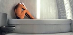 Person in an orange jumpsuit curled up alone in a bare cell